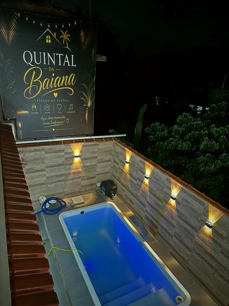 Piscina iluminada de azul com luminárias na parede e o banner do Quintal da Baiana ao fundo